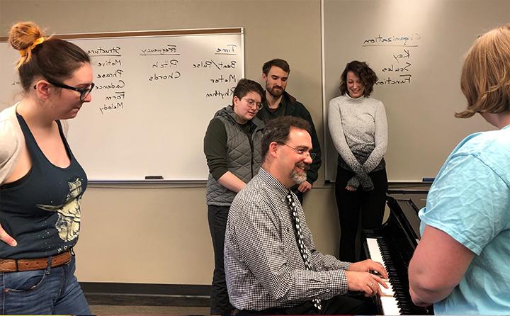 Honors students standing around BW professor playing piano in classroom